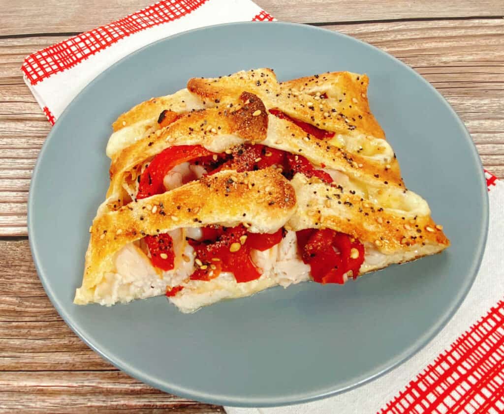 a blue plate sits on a red and white checkered napkin. On the plate is a slice of the braided turkey and red pepper sandwich. Roasted red peppers and turkey peek out from between the braids of pizza dough. The bread is toasted and golden and sprinkled with everything bagel seasoning.