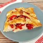 a blue plate sits on a red and white checkered napkin. On the plate is a slice of the braided turkey and red pepper sandwich. Roasted red peppers and turkey peek out from between the braids of pizza dough. The bread is toasted and golden and sprinkled with everything bagel seasoning.