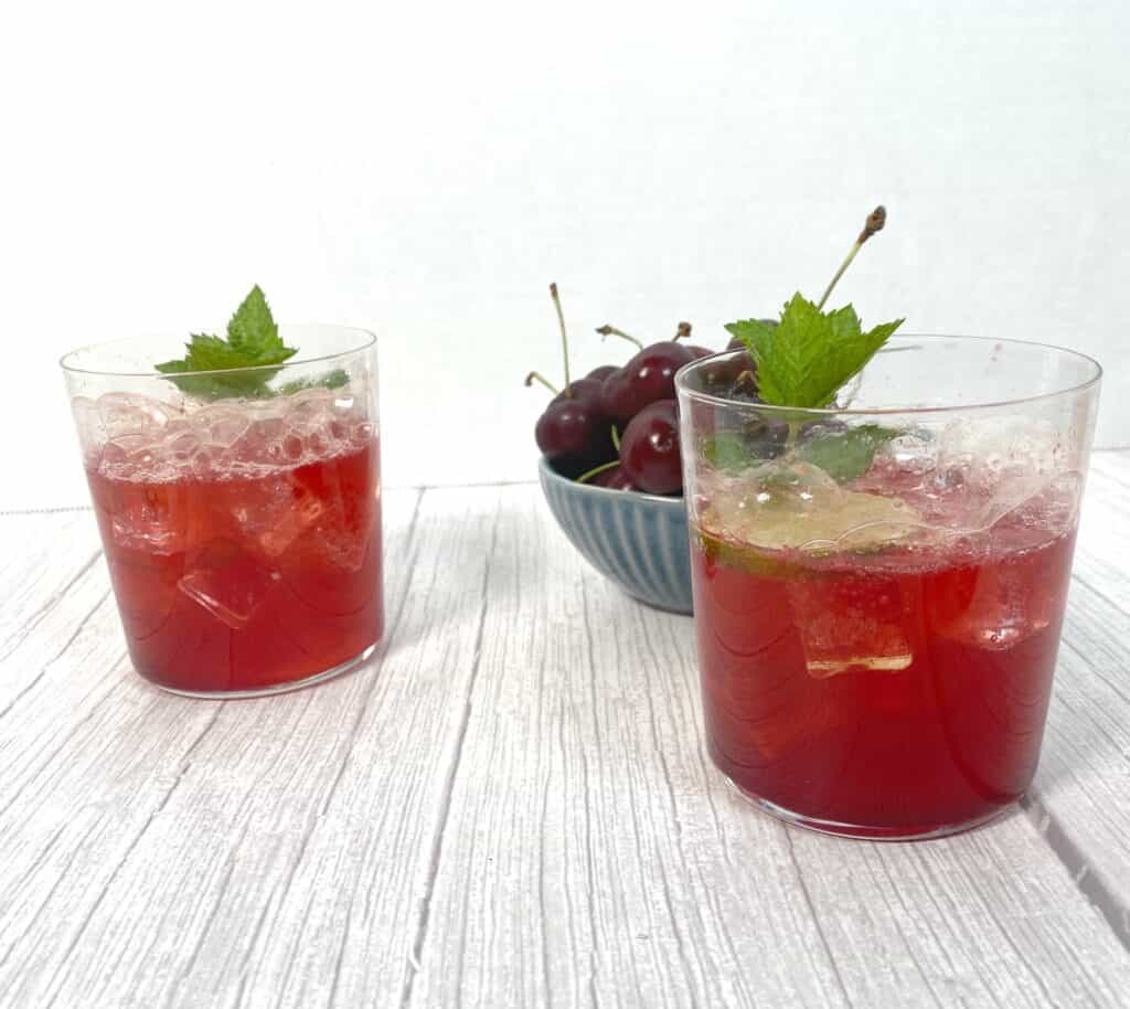 cherry lime bourbon cocktail in glasses on white wood background. bowl of fresh cherries in background
