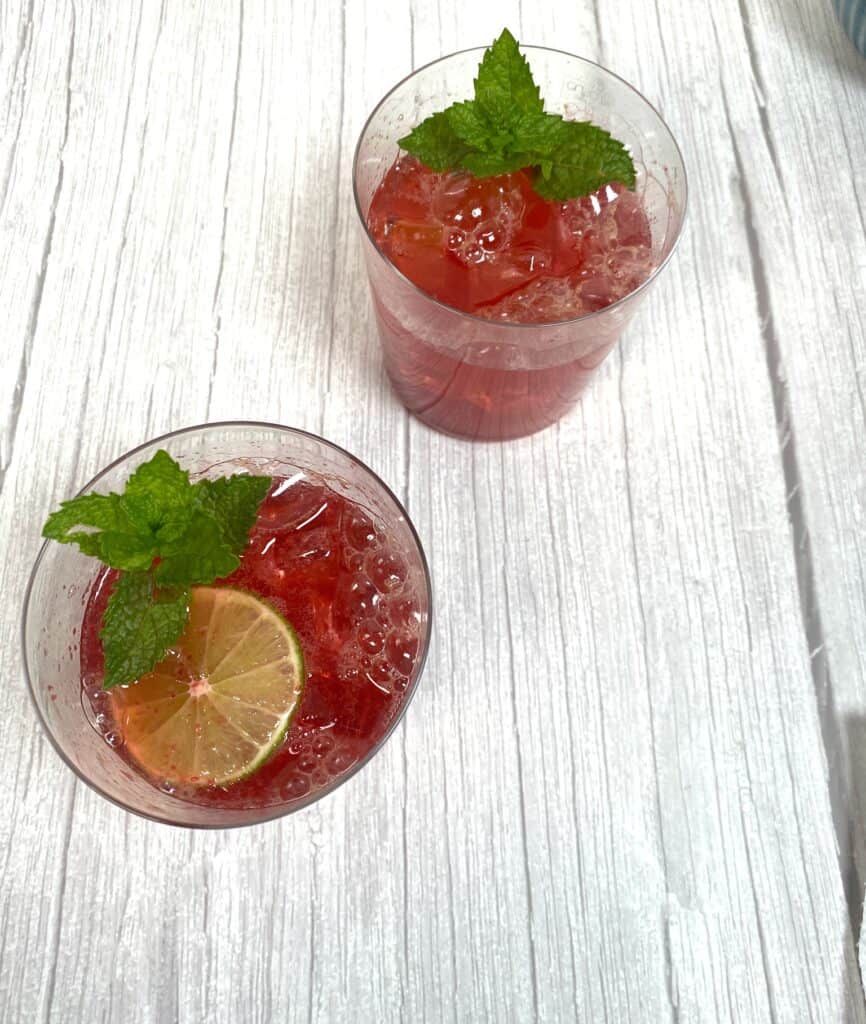cherry lime bourbon cocktail in glass on white wood. drinks are garnished with mint sprigs and lime rounds