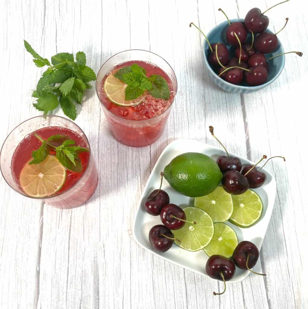 red cocktail made with cherry juice, lime juice and bourbon in glasses. garnished with lime slice and mint. additional cherries and lime slices in image
