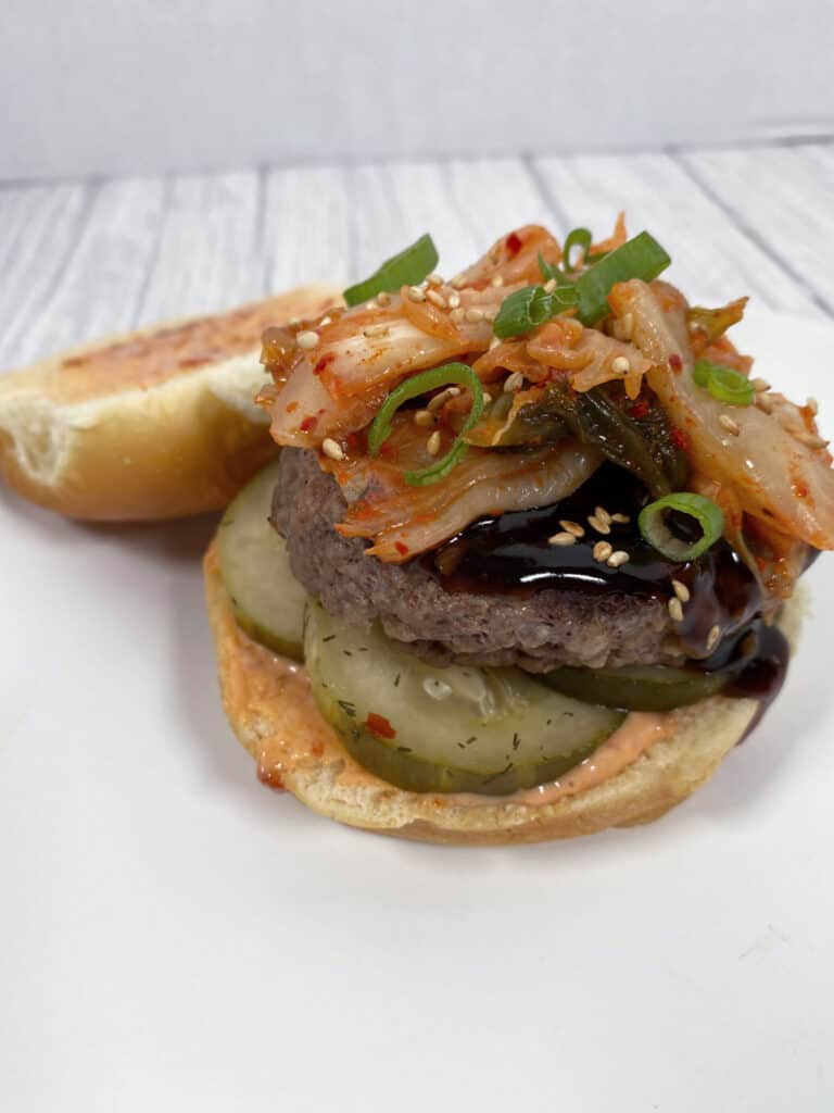 beef burger topped with korean bbq sauce, kimchi and diced green onions. burger is sitting open face to show top bun covered in spicy mayo. 