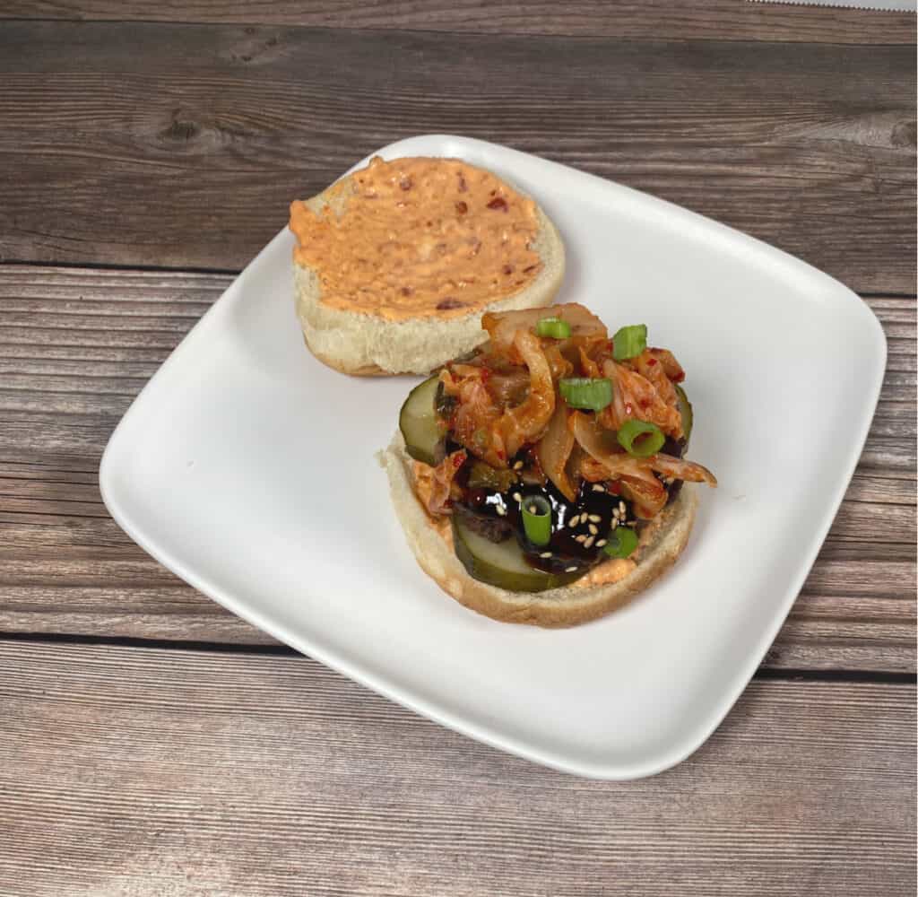 wood background, white plate. Open faced burger sits on white plate. Top half of bun is upside down to show it's slathered with spicy mayo. Burger sits on sliced cucumbers and is topped with korean barbecue sauce, kimchi and diced green onions. 