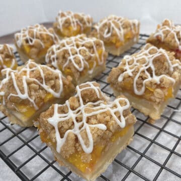 black wire rack with peach crumb bar squares on top. Shortbread crust is visible followed by layer of chopped peaches then topped with a crumble. White glaze zigzags on top of each bar.
