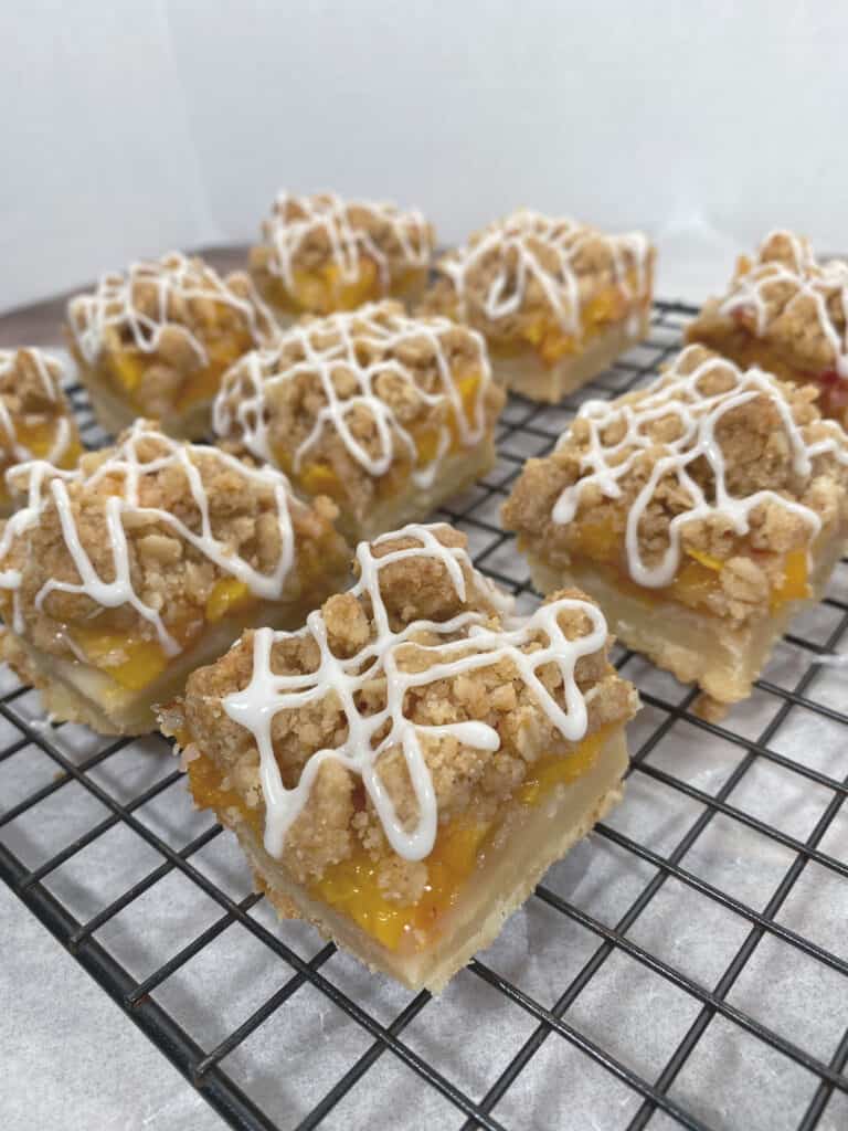 black wire rack with peach crumb bar squares on top. Shortbread crust is visible followed by layer of chopped peaches then topped with a crumble. White glaze zigzags on top of each bar.