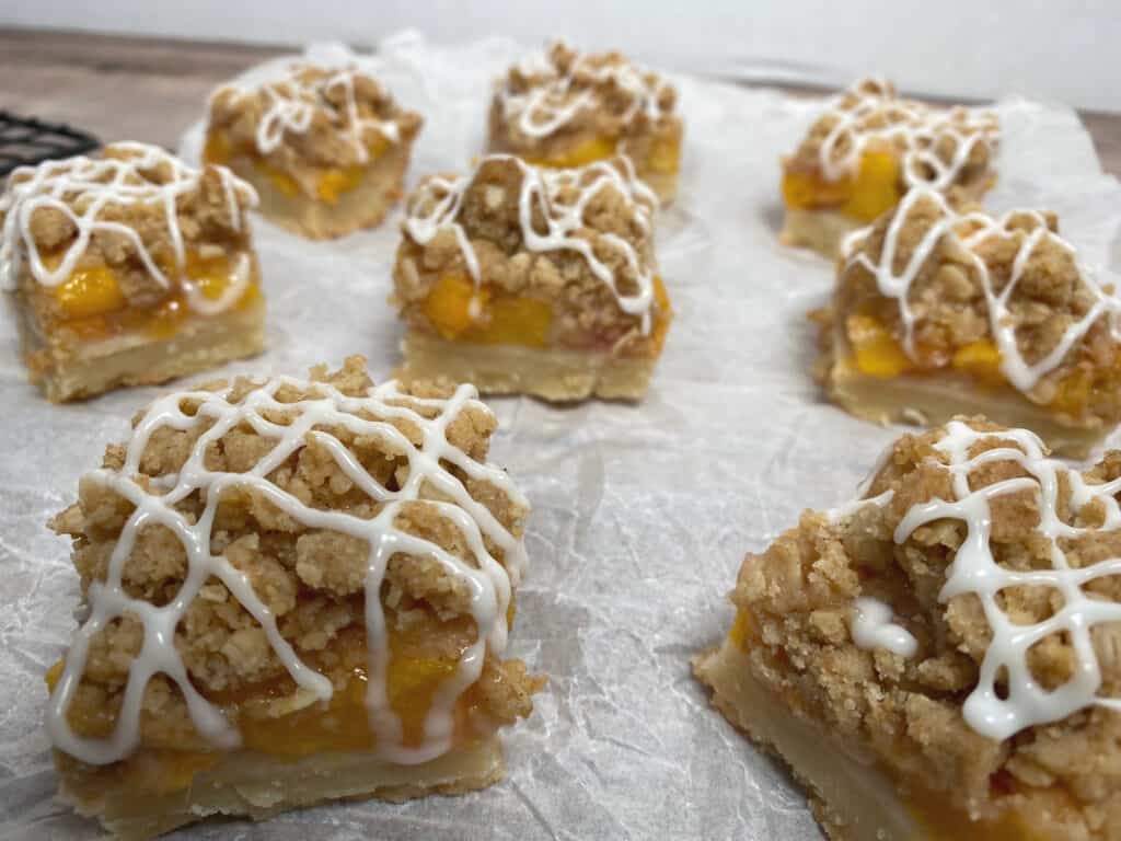 white parchment paper background. Peach crumb bar with visible layers of peaches and oat crumble, topped with zigzag vanilla glaze.