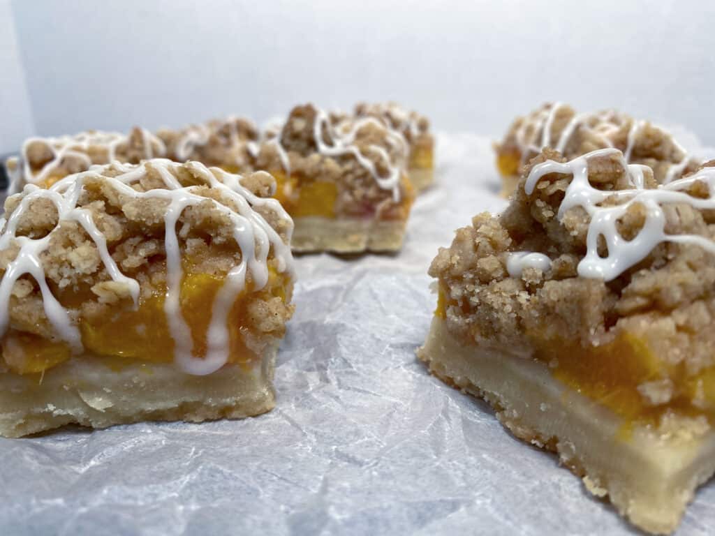 peach crumb bar squares on top. Shortbread crust is visible followed by layer of chopped peaches then topped with a crumble. White glaze zigzags on top of each bar.