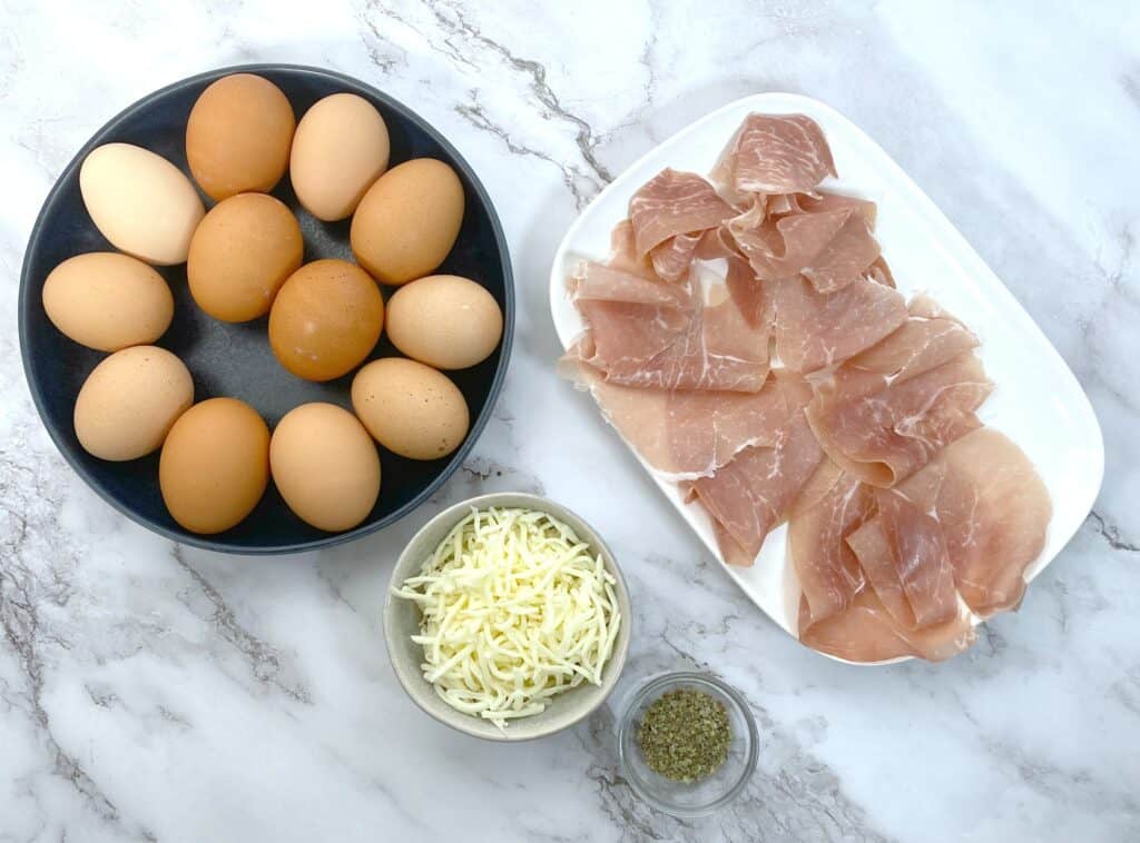 Ingredients for prosciutto egg cups on a marble background - a bowl of farm-fresh brown eggs sits on the left. On the right is a small white platter layered with thin slices of prosciutto. Below that is a bowl of shredded mozzarella cheese and a small glass container of italian seasoning. 