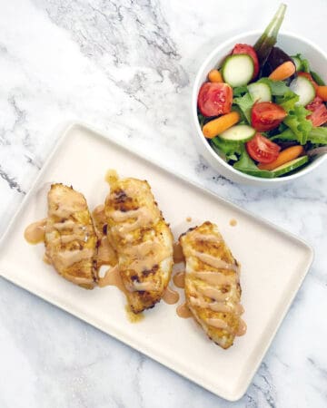 baked chicken breasts covered with spicy bbq drizzle next to a garden salad on a marble background