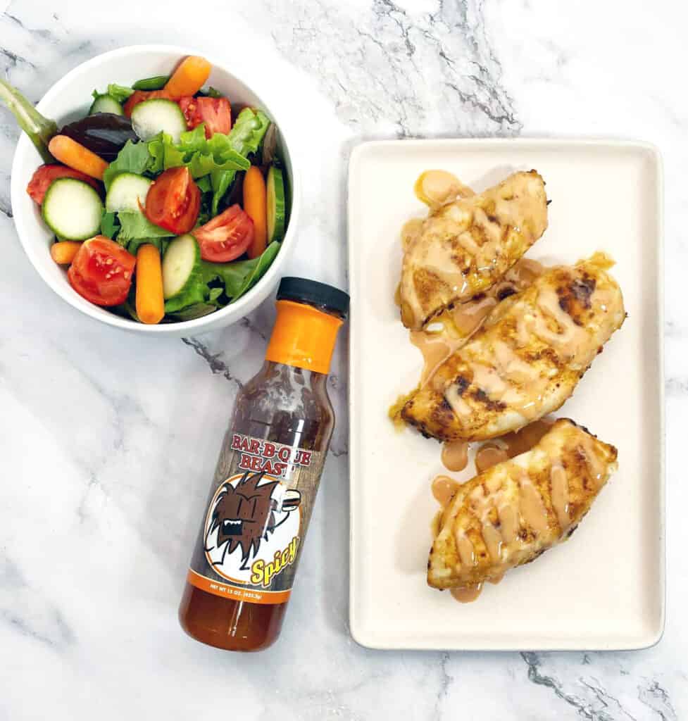 baked chicken breasts on a rectangle platter. Chicken breasts are drizzled with spicy BBQ sauce. Sauce bottle is pictured as is a garden salad on a marble background