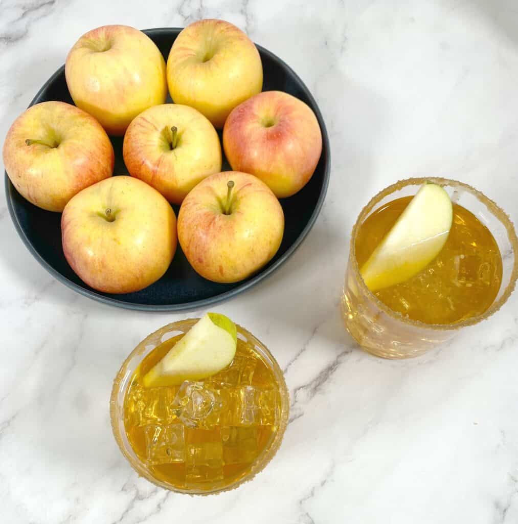 caramel apple whiskey sour cocktails with bowl of apples behind them