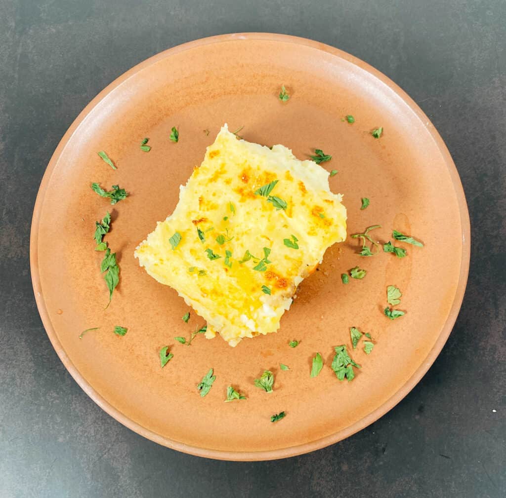 Slice of shepherd’s pie on a copper colored plate. Cheesy mashed potatoes are layered on top of a meat and veggie filling. The plate is garnished with parsley. 