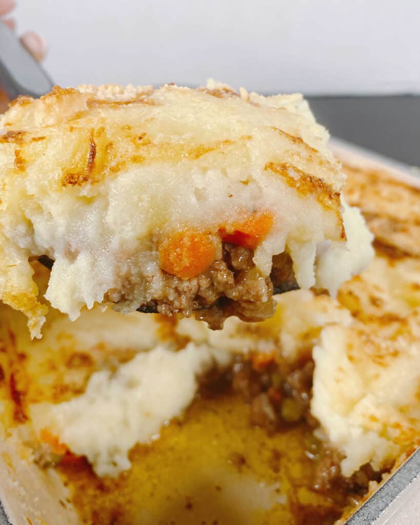 A spatula lifts up a serving of shepherd’s pie from the baking dish it cooked in. Cheesy mashed potatoes are topping a meat and veggie mixture. 