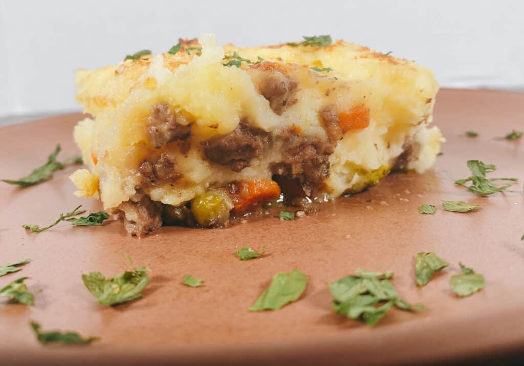 Slice of shepherd’s pie on a copper colored plate. Cheesy mashed potatoes are layered on top of a meat and veggie filling. The plate is garnished with parsley. 