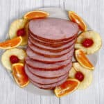 Ham, coated in a brown sugar and mountain dew glaze, sits sliced on a platter, surrounded by pineapple rings, maraschino cherries and orange wedges.