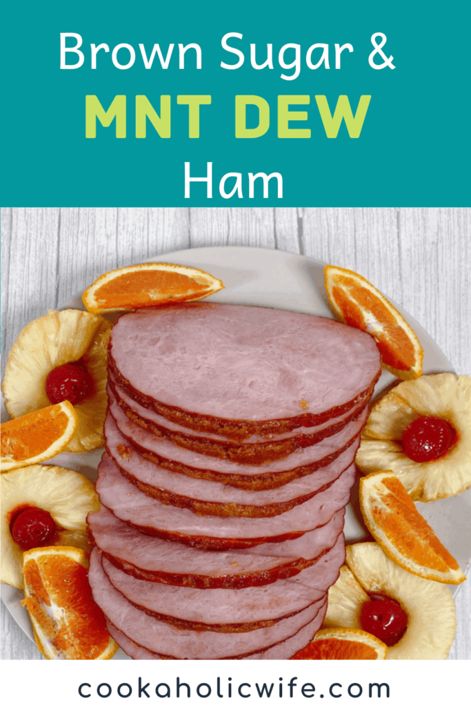 Ham, coated in a brown sugar and mountain dew glaze, sits sliced on a platter, surrounded by pineapple rings, maraschino cherries and orange wedges. 