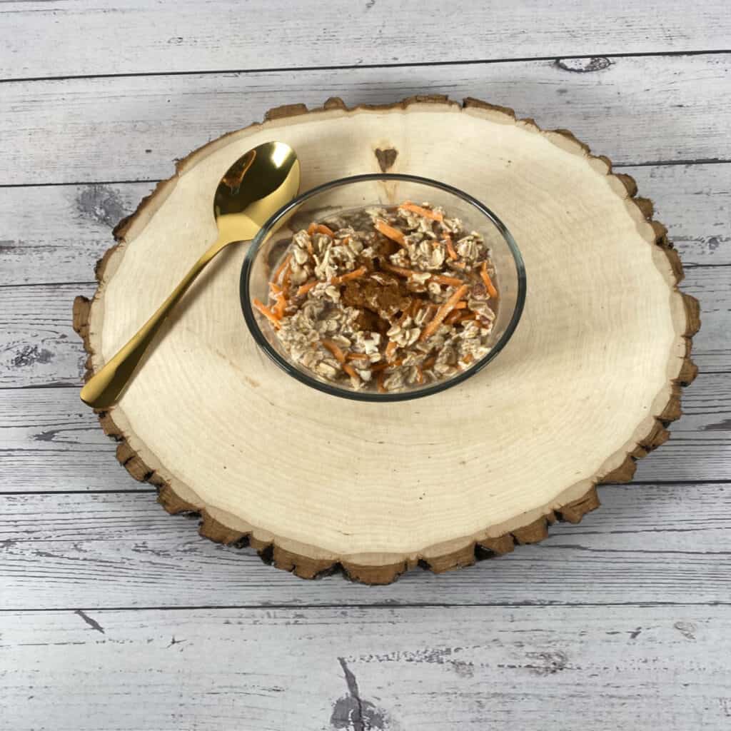 A bowl of carrot cake overnight oats sits on top of a wooden log with a golden spoon to the left. 