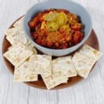 a blue bowl filled with beef chili, topped with shredded cheese and pickled jalapenos sits on a terra cotta colored plate surrounded with buttered saltine crackers