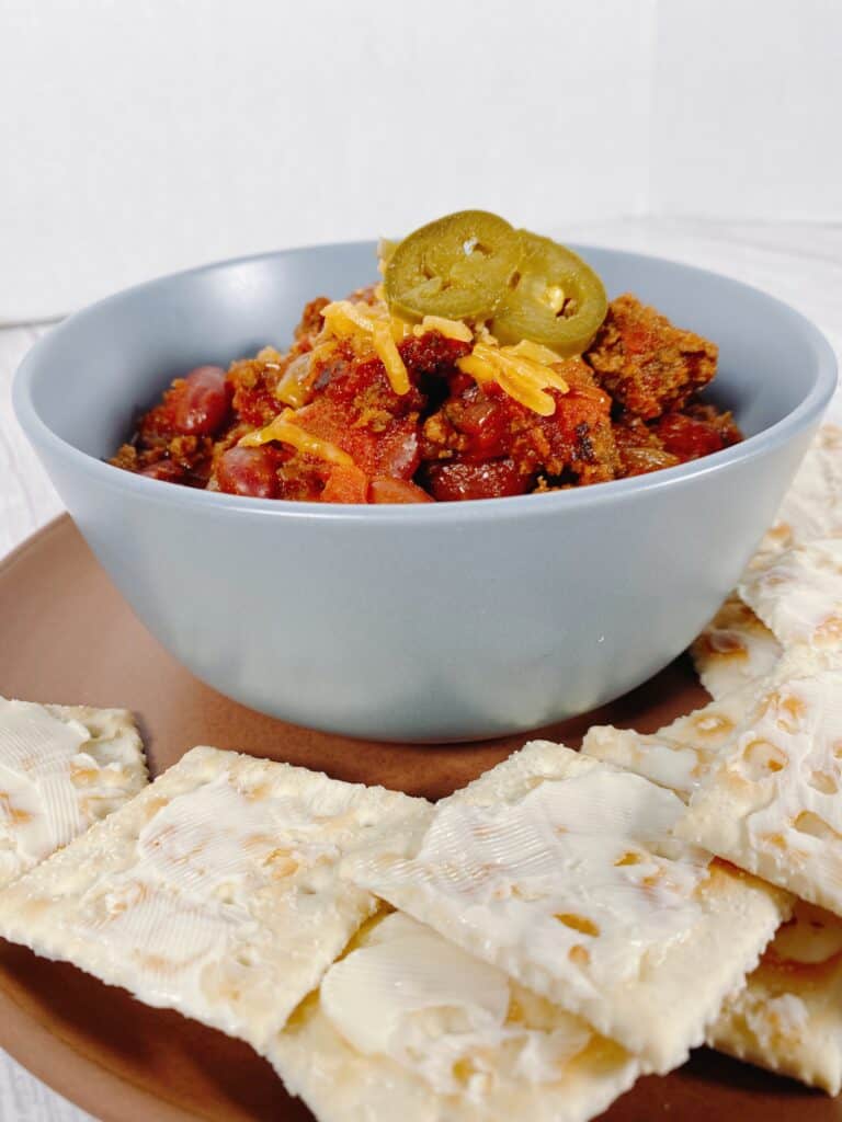 Light blue bowl filled with beef and bean chili, topped with shredded cheddar cheese and pickled jalapeno slices. The bowl sits on top of a terra cotta colored plate that is full of buttered saltine crackers. 