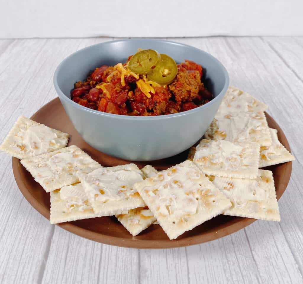 Light blue bowl filled with beef and bean chili, topped with shredded cheddar cheese and pickled jalapeno slices. The bowl sits on top of a terra cotta colored plate that is full of buttered saltine crackers.