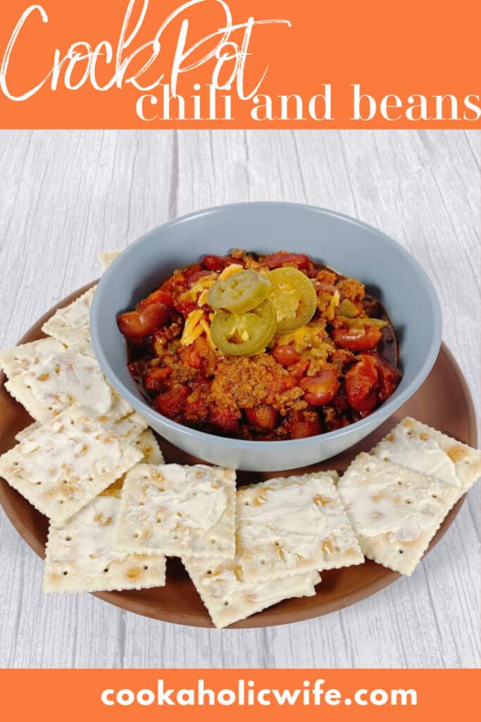Light blue bowl filled with beef and bean chili, topped with shredded cheddar cheese and pickled jalapeno slices. The bowl sits on top of a terra cotta colored plate that is full of buttered saltine crackers. 