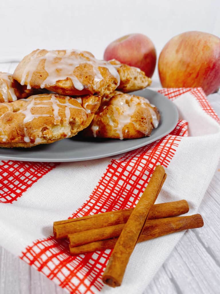 Air fryer apple fritters sit on a blue plate with a red and white checkered napkin. Apples and cinnamon sticks surround them.