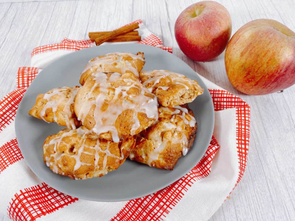 Air fryer apple fritters sit on a blue plate with a red and white checkered napkin. Apples and cinnamon sticks surround them.