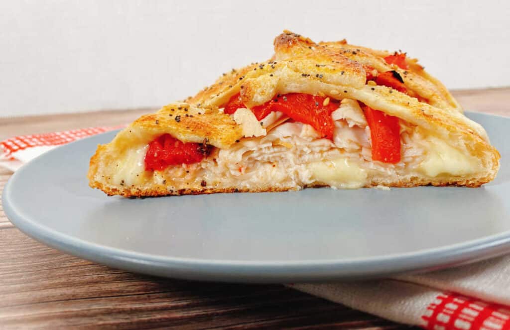 front view of a slice of the braided turkey and red pepper sandwich. A red and white checkered napkin sits on a wooden background. On top of the napkin is a blue plate, with the sandwich on top. Cheese melts out from the bottom of the sandwich, followed by layers of turkey and roasted red peppers. The top of the braid is golden brown and sprinkled with everything bagel seasoning. 