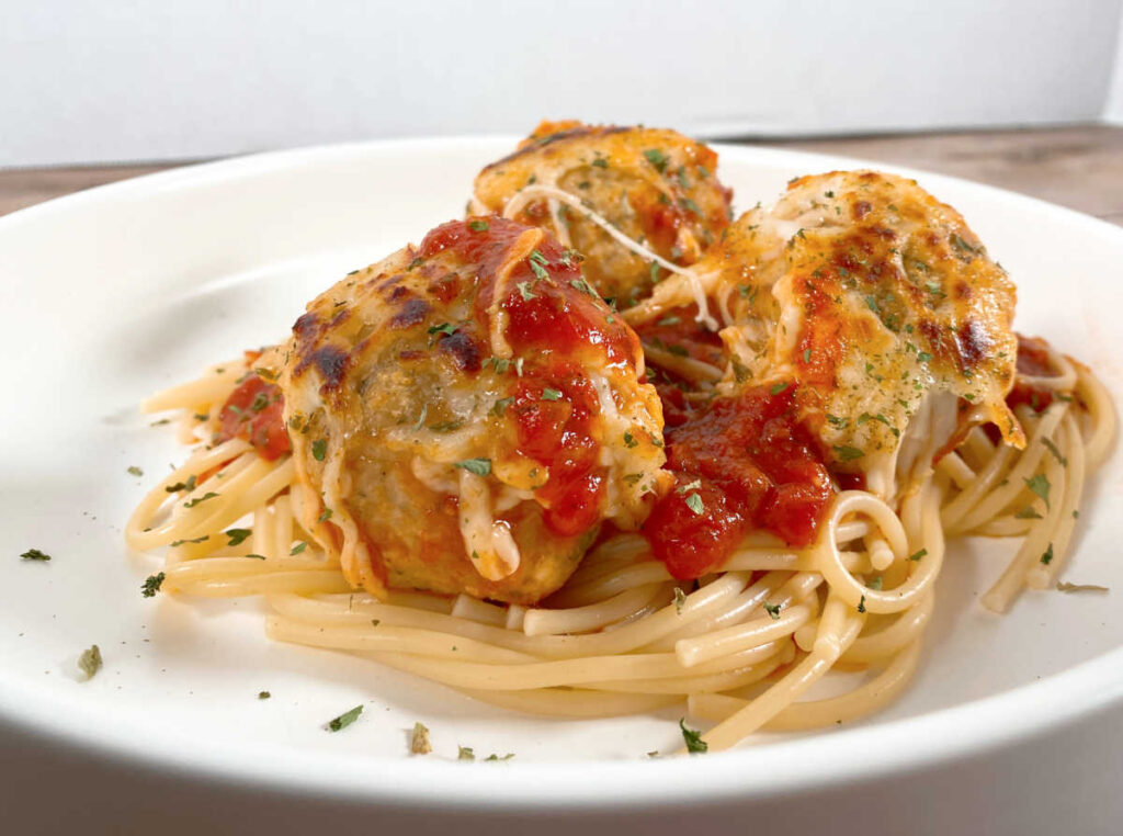 Cheese covered meatballs sit on top of pasta in a white pasta dish. 