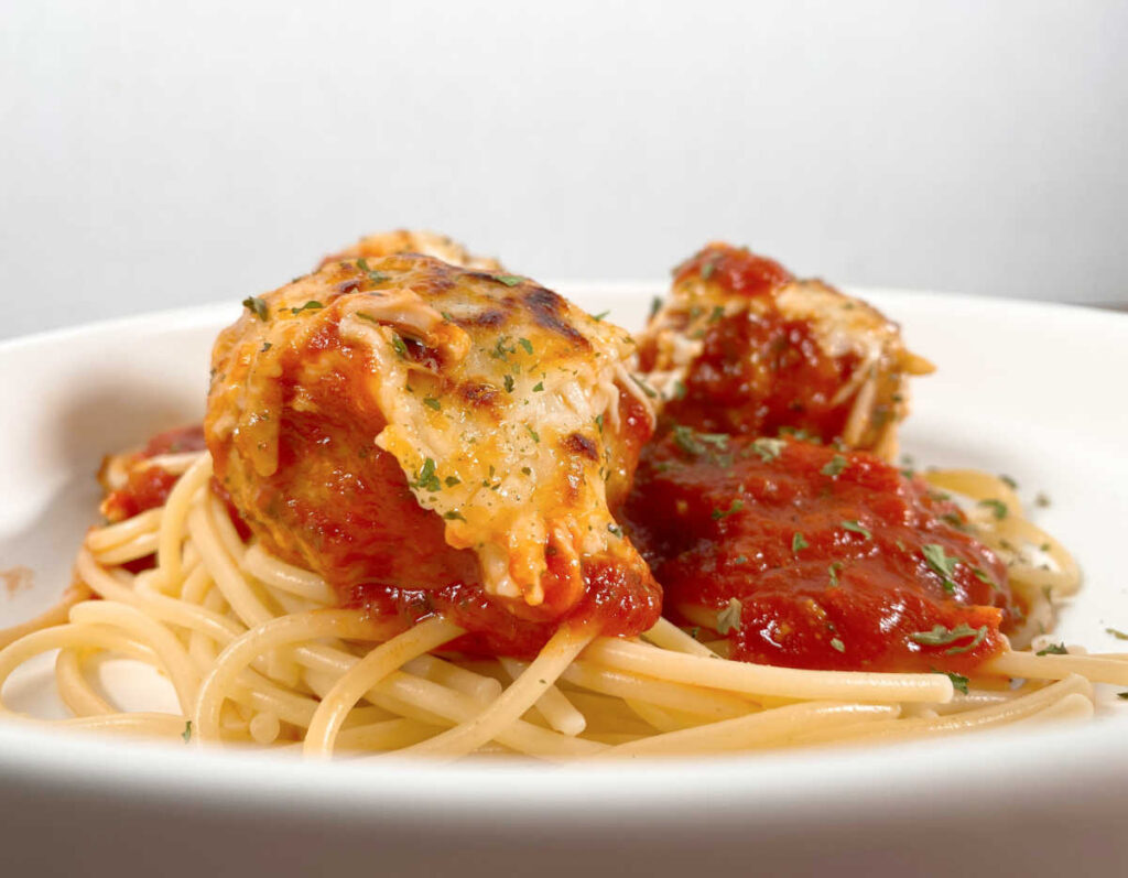 Cheese and sauce covered meatballs sit over a bed of pasta in a shallow white pasta dish. 