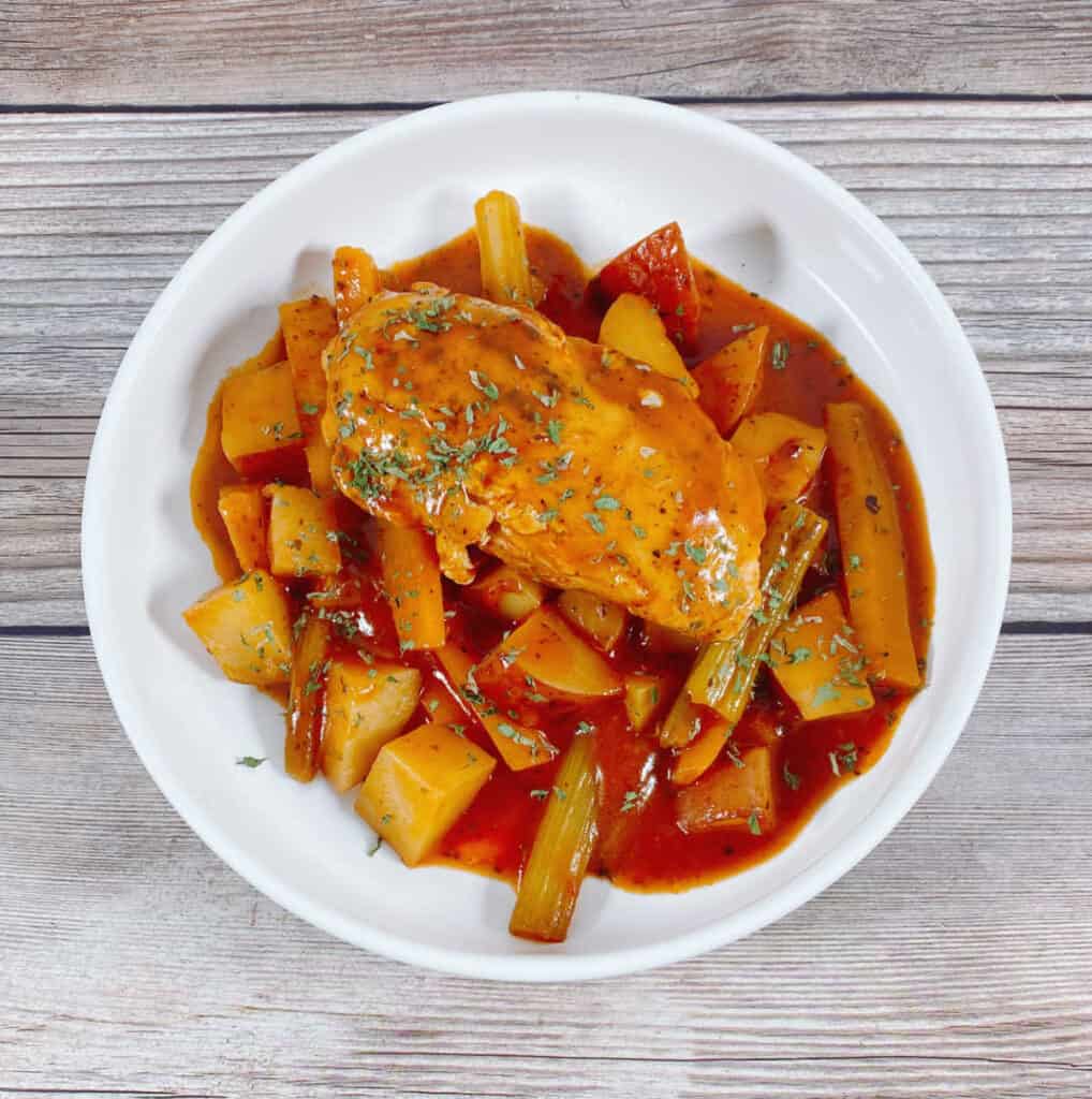 on a wooden background with a tan napkin sits a white bowl of braised chicken; chicken, carrots, celery and potatoes in a tomato broth.