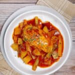 on a wooden background with a tan napkin sits a white bowl of braised chicken; chicken, carrots, celery and potatoes in a tomato broth.