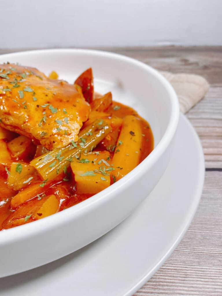 on a wooden background with a tan napkin sits a white bowl of braised chicken; chicken, carrots, celery and potatoes in a tomato broth.