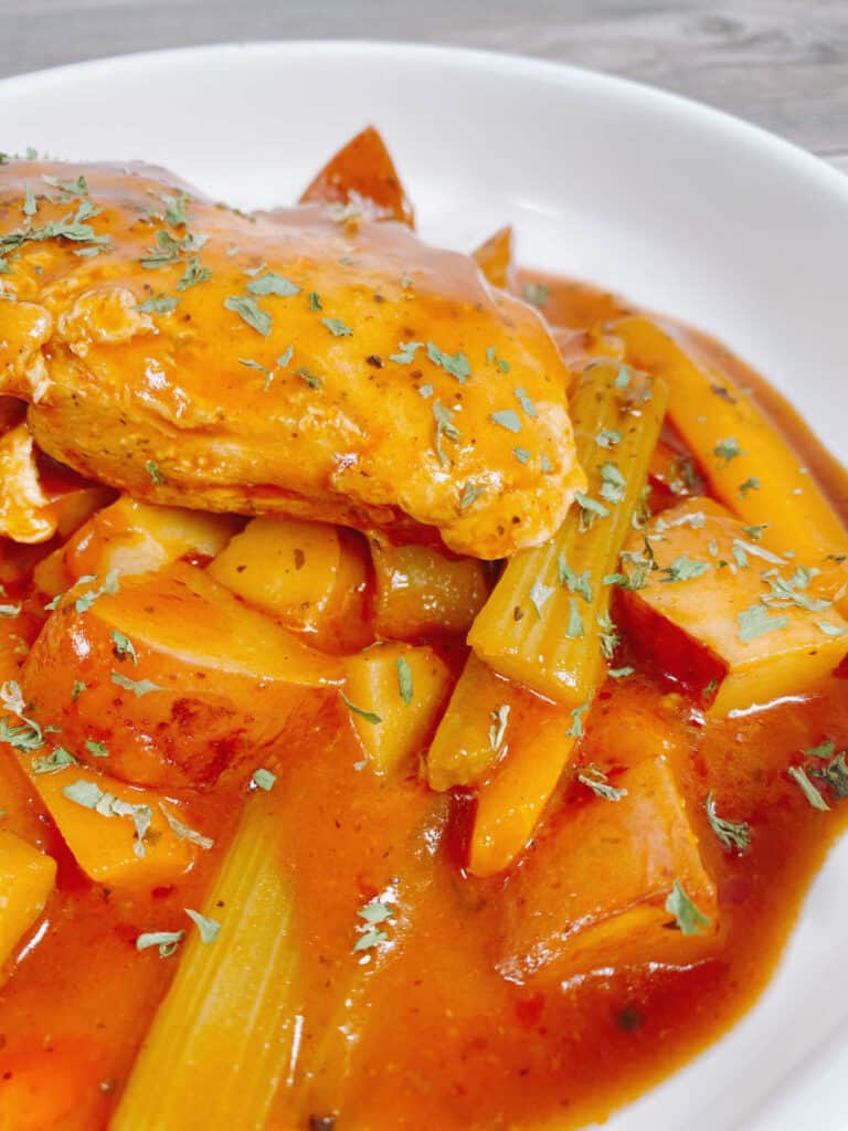 on a wooden background with a tan napkin sits a white bowl of braised chicken; chicken, carrots, celery and potatoes in a tomato broth.