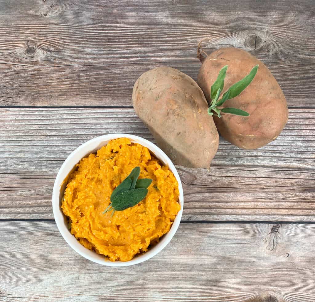 mashed sweet potatoes in a white bowl, topped with fresh sage leaves. Beside it, raw sweet potatoes. 