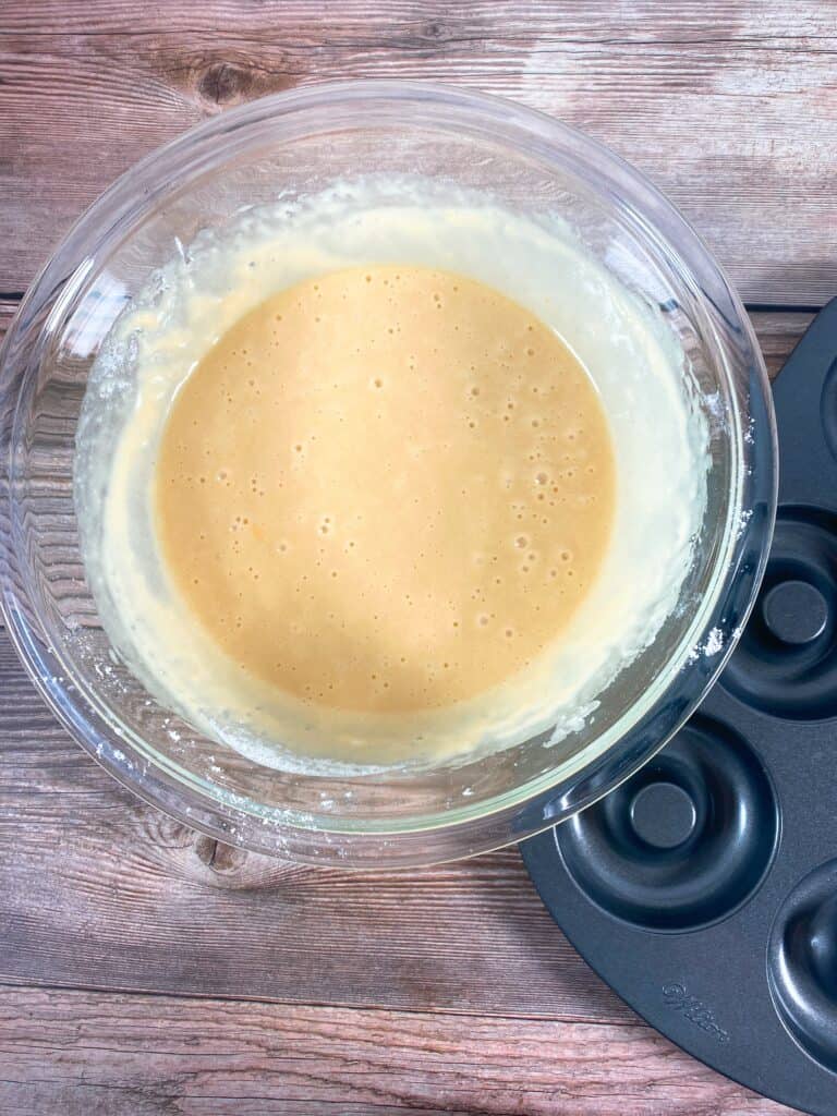 batter for baked cinnamon sugar donuts sits in a glass bowl on a wooden background. on the side of the bowl is a donut pan. 