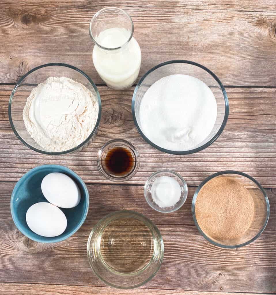 ingredients for baked cinnnamon sugar donuts  in bowls on a wooden background - all purpose flour, sugar, buttermilk, vanilla extra, salt, baking powder, eggs, canola oil and a cinnamon sugar mixture for the coating.