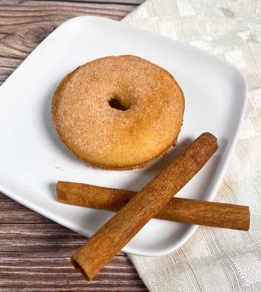 baked donut coated in cinnamon sugar sits on a white plate on a wooden background. two cinnamon sticks are criss crossed in front of the donut.