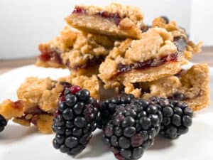 stacked up blackberry oat crumble bars sit on a white plate with fresh blackberry sprinkled around them