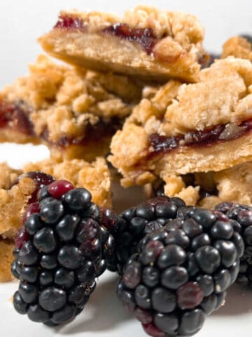 stacked up blackberry oat crumble bars sit on a white plate with fresh blackberry sprinkled around them