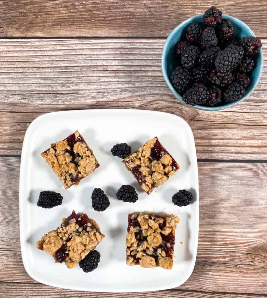 A white plate of blackberry crumble bars with fresh blackberries sits on a wooden background. To the left is a bowl of fresh blackberries 