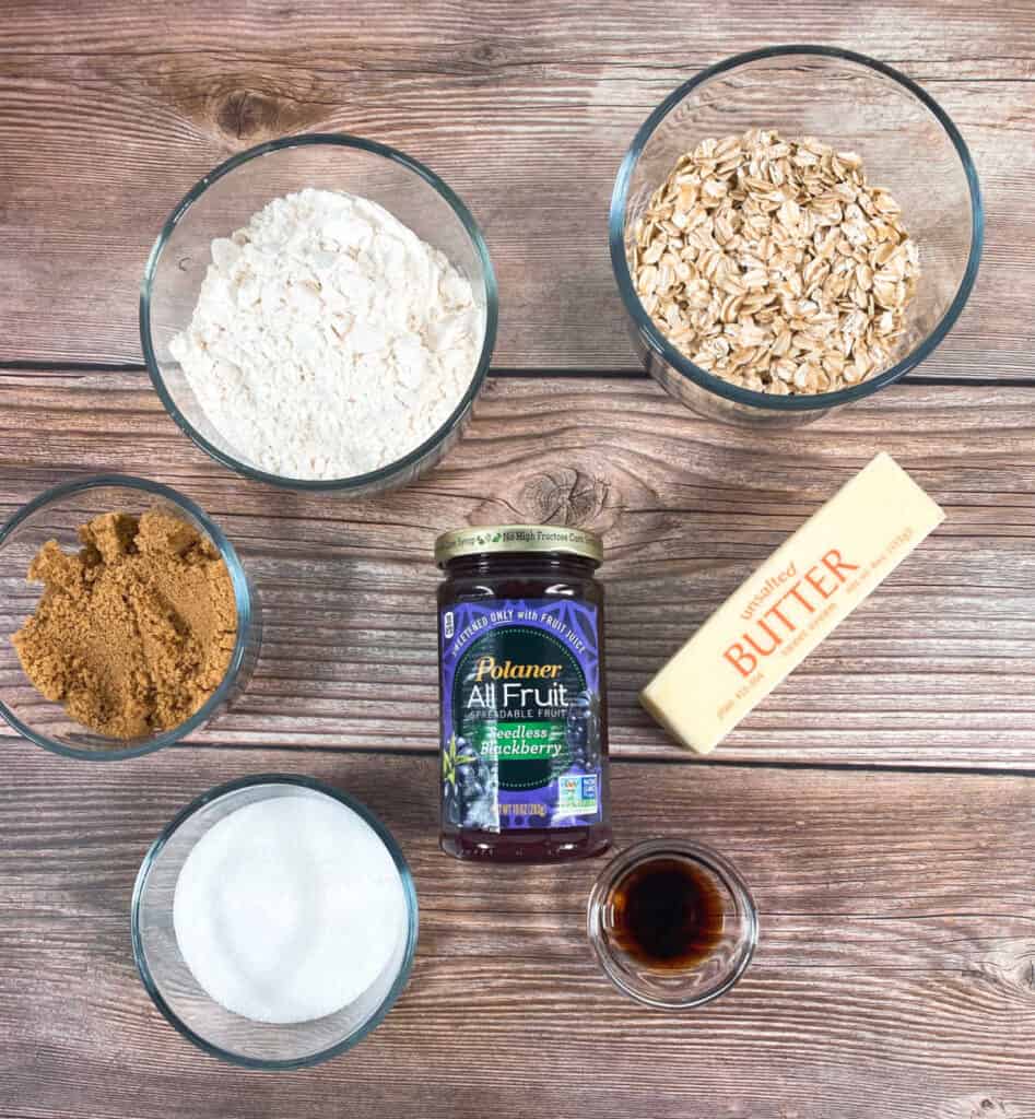 ingredients for blackberry oat crumble bars on a wooden background - all purpose flour, brown sugar, granulated sugar, old fashioned oats, butter, vanilla and blackberry preserves 