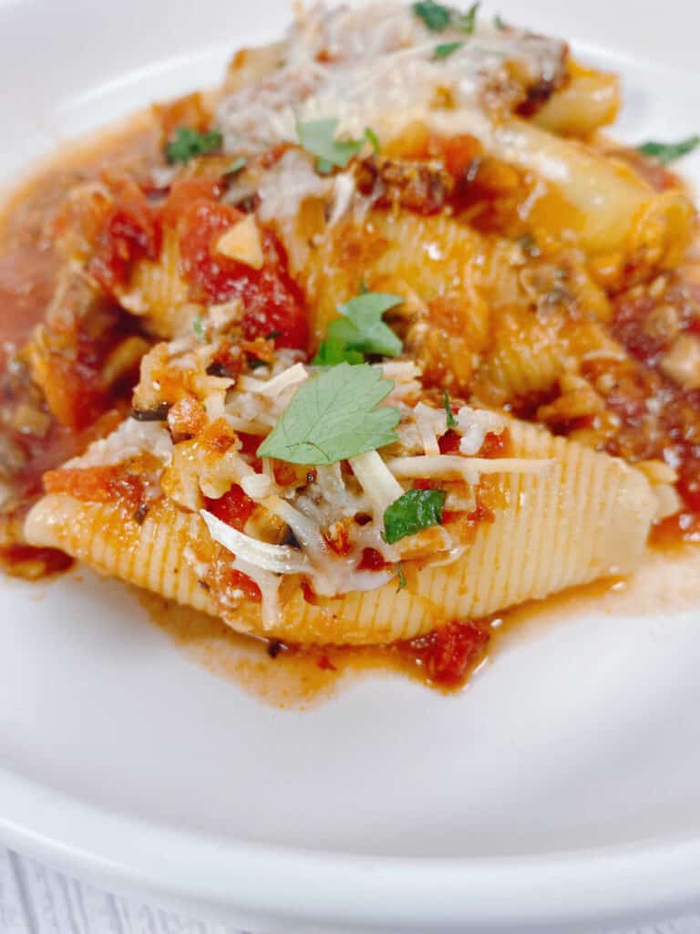 four cheesy vegetarian cheesy stuffed shells line up in a row on a white plate. The shells are topped with mozzarella cheese and fresh parsley. 