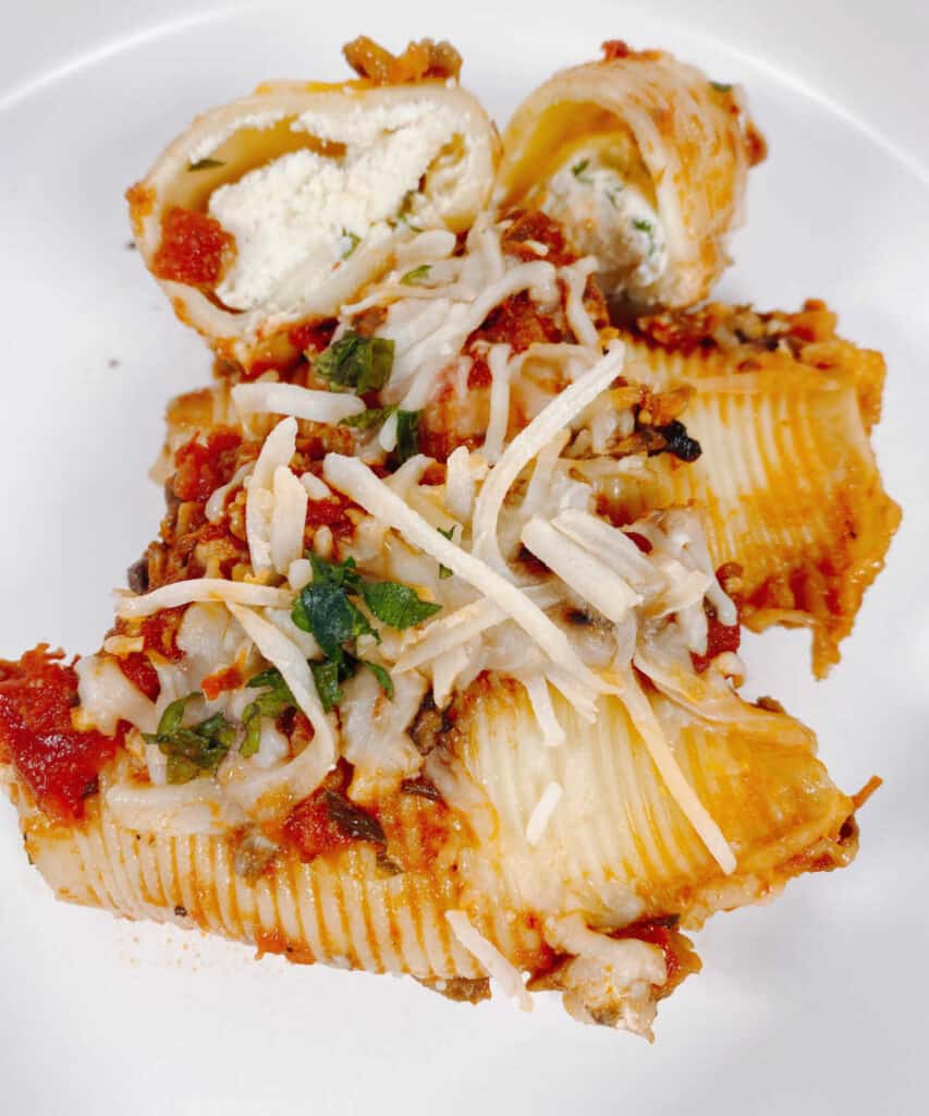 Cheesy vegetarian stuffed shells are on a white plate. One of the shells is cut open to show the cheese filling inside. the others are topped with mozzarella cheese and parsley. 