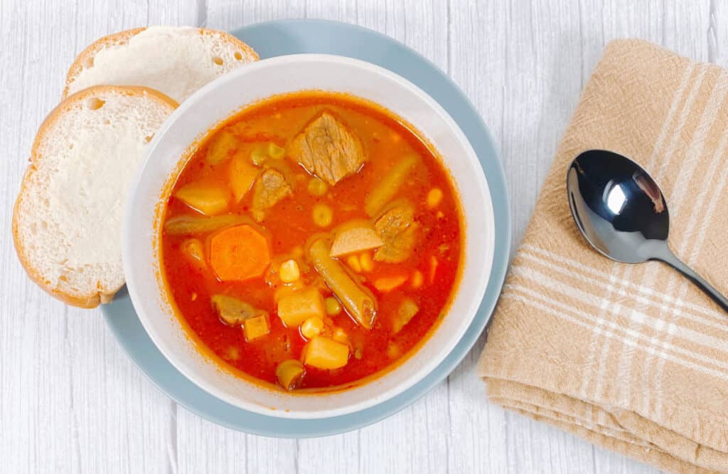 Photo is taken looking down on the soup. There is a white wooden background. On the left is a blue plate with stacked slices of buttered bread. Next to the bread is a tomato based Grandma's beef and vegetable soup. To the right of the bread is a folded, tan, checkered napkin with a spoon on top. 