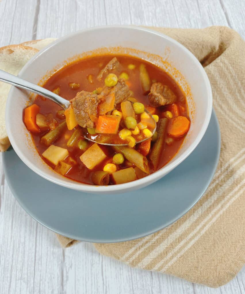 top down view - looking a spoon lifting the ingredients of a tomato based beef and vegetable soup. Under the bowl of soup is a blue plate and a tan checkered napkin.