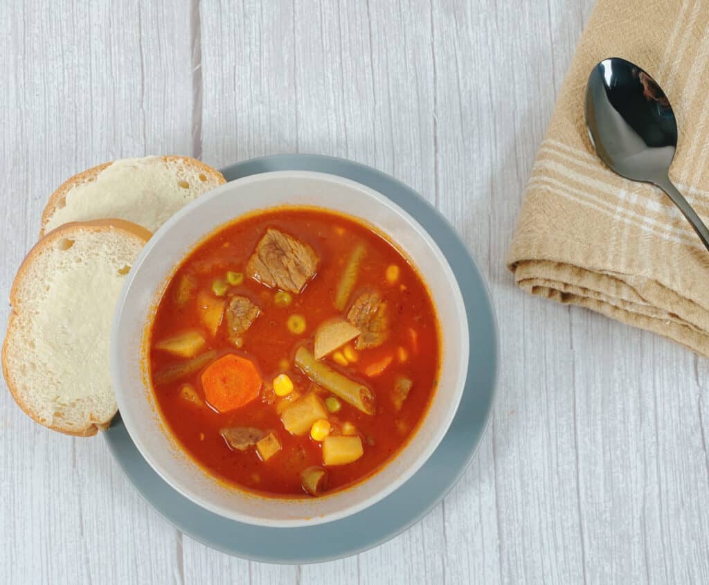 Image is looking down at the bowl of Grandma's Beef and Vegetable Soup. White wooden background with a blue plate on the left. Left side of the plate is stacked slices of buttered bread. Next to that, in a white bowl is the beef and vegetable soup. On the right side of the image is a folded tan, checkered napkin with a spoon on top. 