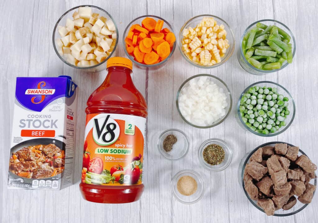 ingredients for grandma's beef and vegetable soup sit on a white wooden background. Top row - diced potatoes, carrots, corn and green beans in glass dishes. Bottom row - carton of beef stock, container of V8 spicy vegetable juice, bowl of diced onions, bowl of frozen peas, bowl of beef cubes and small containers holding salt, pepper, garlic powder and dried oregano. 