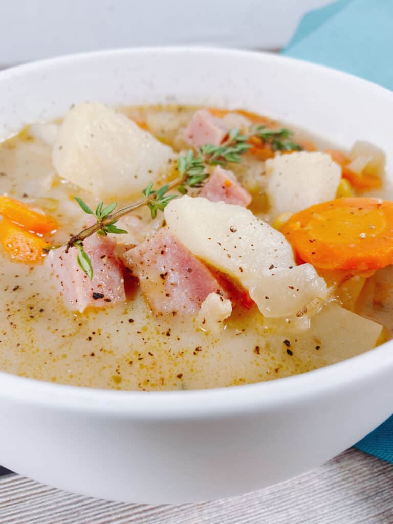 Close up of creamy ham and potato soup in a white bowl. Pieces of ham, potato and carrot rest on top of the broth, seasoned with salt and pepper. A sprig of thyme sits on top of the bowl. 