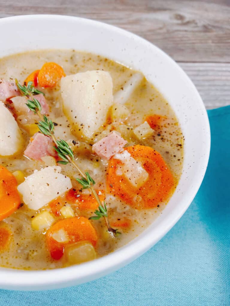 A white bowl of creamy ham and potato soup covers nearly all of the image. In the bottom corner is a turquoise napkin. The bowl of soup is full of pieces of potato, ham, carrot and corn. Salt and pepper season the dish, with a sprig of fresh thyme on top. 